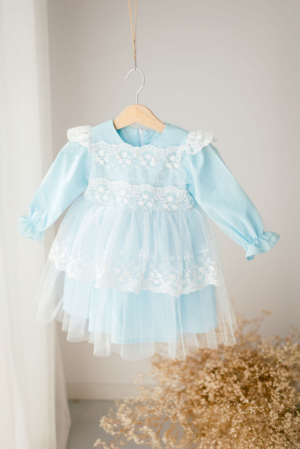 A light blue dress with white lace trim is hanging on a wooden hanger against a white wall.