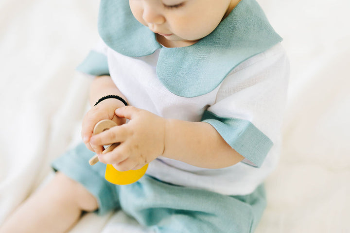 A baby wearing a white shirt with a blue collar and light blue shorts is sitting on a white surface, holding a yellow object in their hands.