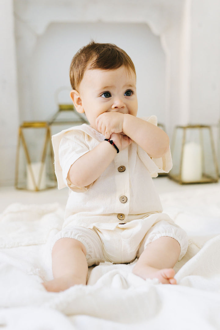 A baby in a white outfit sits on a white blanket, holding their hands to their mouth.