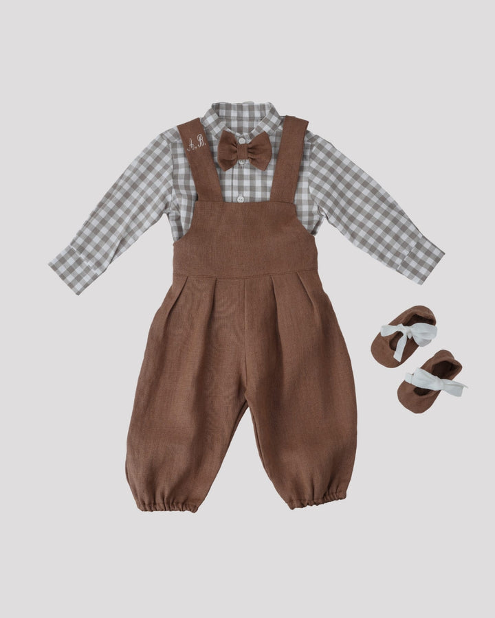 A set of children's clothing consisting of a plaid shirt, brown overalls, and a bow tie, along with a pair of brown shoes.