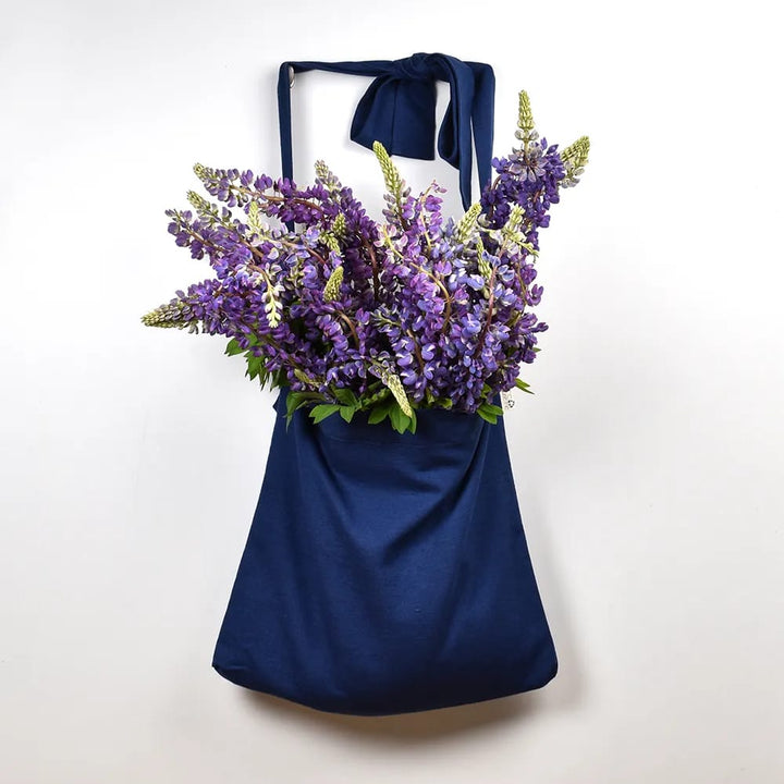 A blue tote bag is hanging against a white wall, filled with a bouquet of purple flowers.