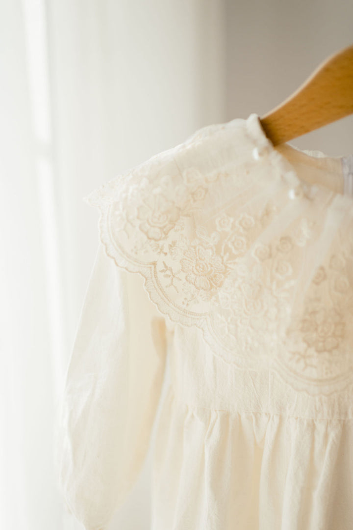 A white lace dress with a scalloped collar is hanging on a wooden hanger against a white background.