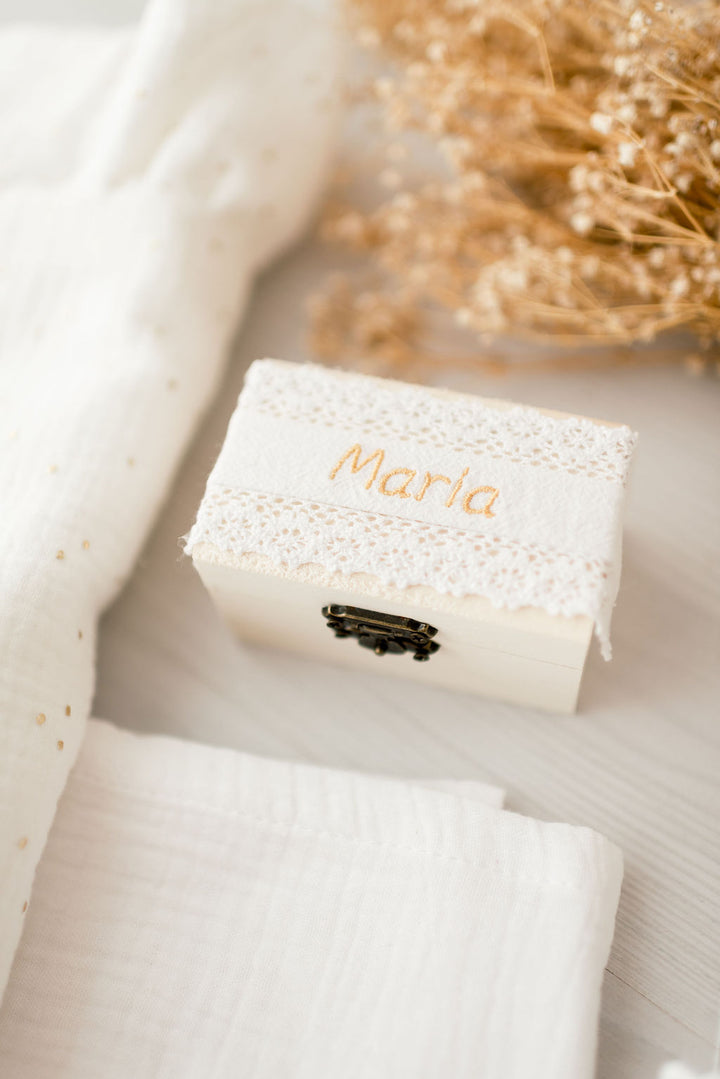 A small white box with a lace-like cover and the name "Marie" written on it, placed on a white surface next to some dried flowers.
