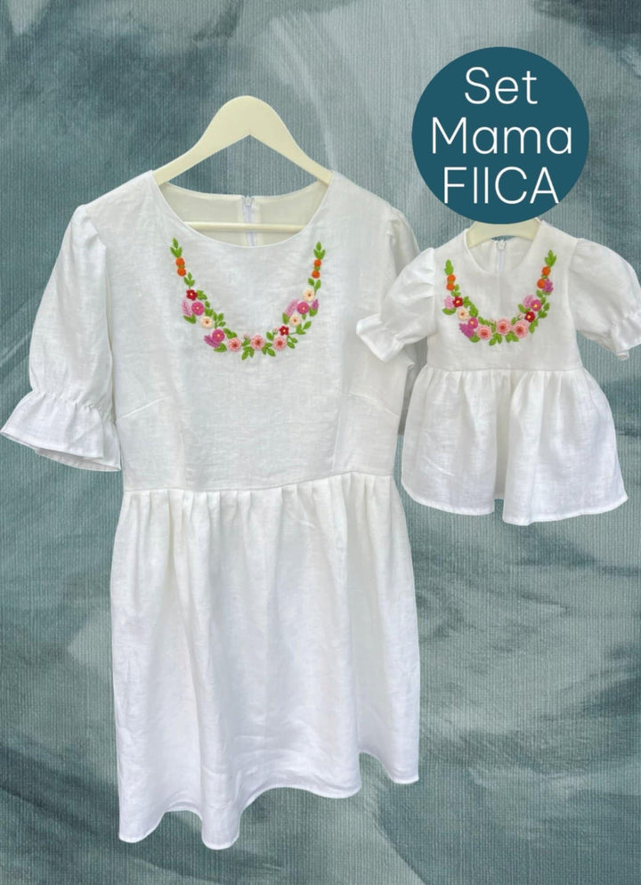a white dress with a floral embroidered design, hanging on a hanger. The dress appears to be a size smaller, as indicated by the smaller size of the doll in the image.
