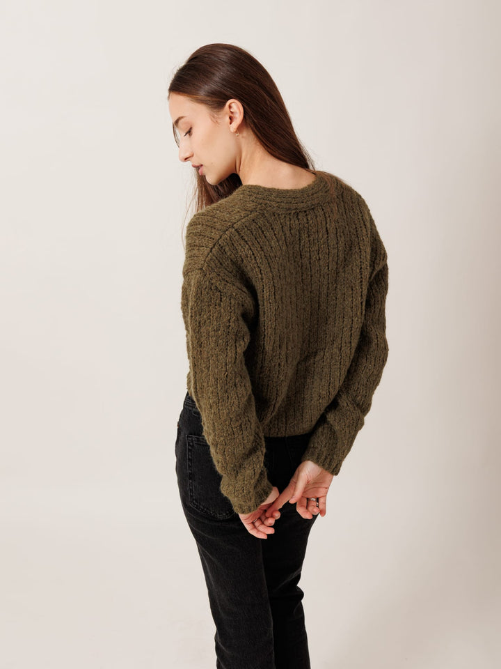 A woman wearing a green sweater stands with her hands behind her back, looking down.