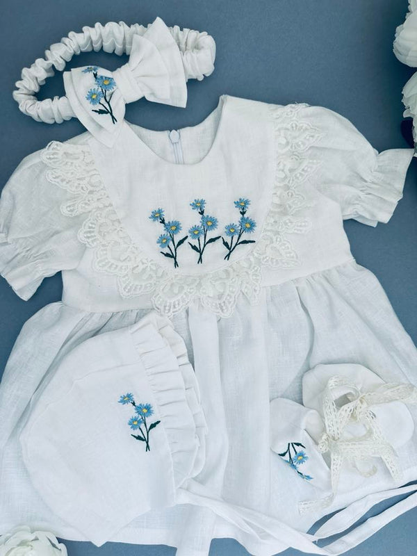 A white dress with blue flowers embroidered on it, along with a matching headband and bonnet, all laid out on a blue background.
