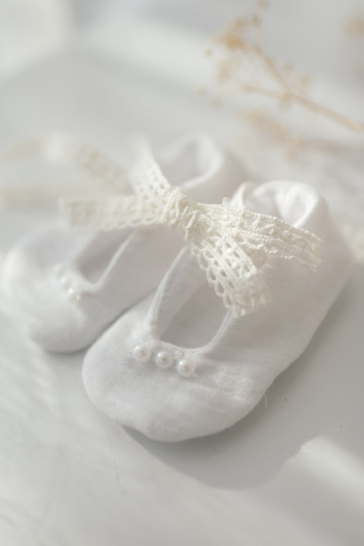 A pair of white baby shoes with a delicate lace bow on top, resting on a white surface.