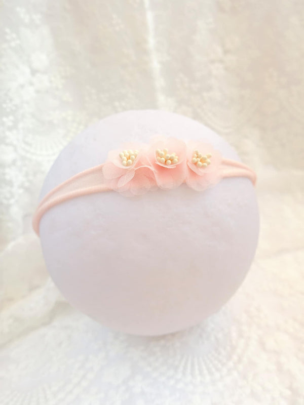 A delicate pink flower headband with three petals is displayed against a white lace background.