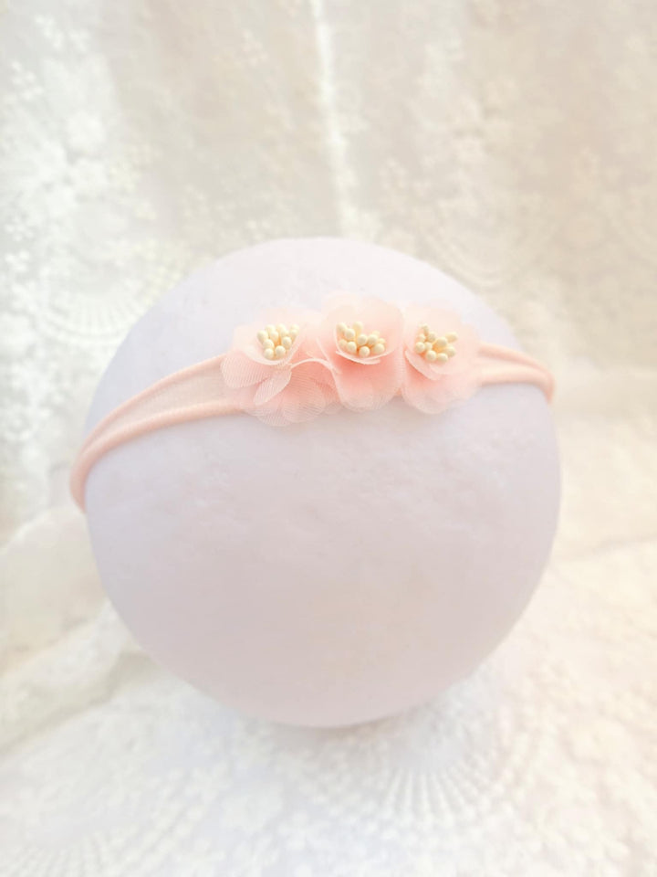 A delicate pink flower headband with three petals is displayed against a white lace background.