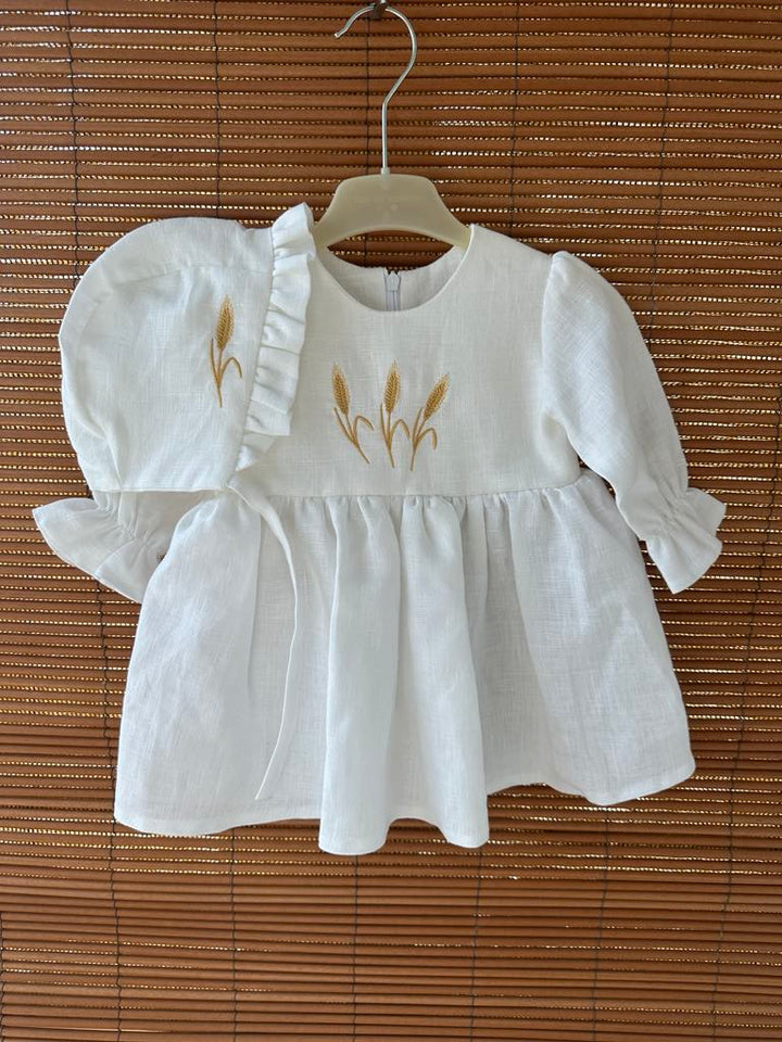 A white dress with gold embroidered wheat stalks on the front, hanging on a wooden hanger.