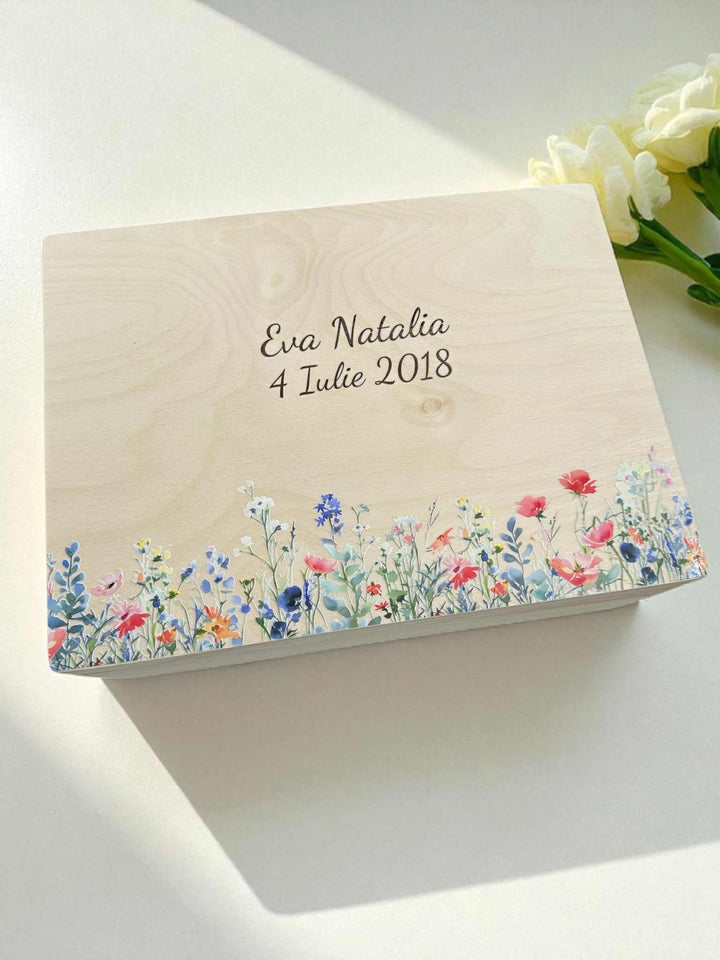 A wooden box with a floral design and the text " Eva Natalie 4 July 2018" printed on it, placed on a white surface next to a bouquet of white flowers.