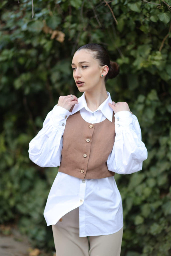 A woman wearing a brown vest, white shirt, and beige pants stands in front of a lush green background.