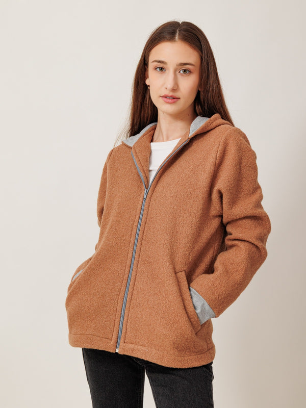 A young woman with long brown hair wearing a brown hooded jacket stands against a plain white background.