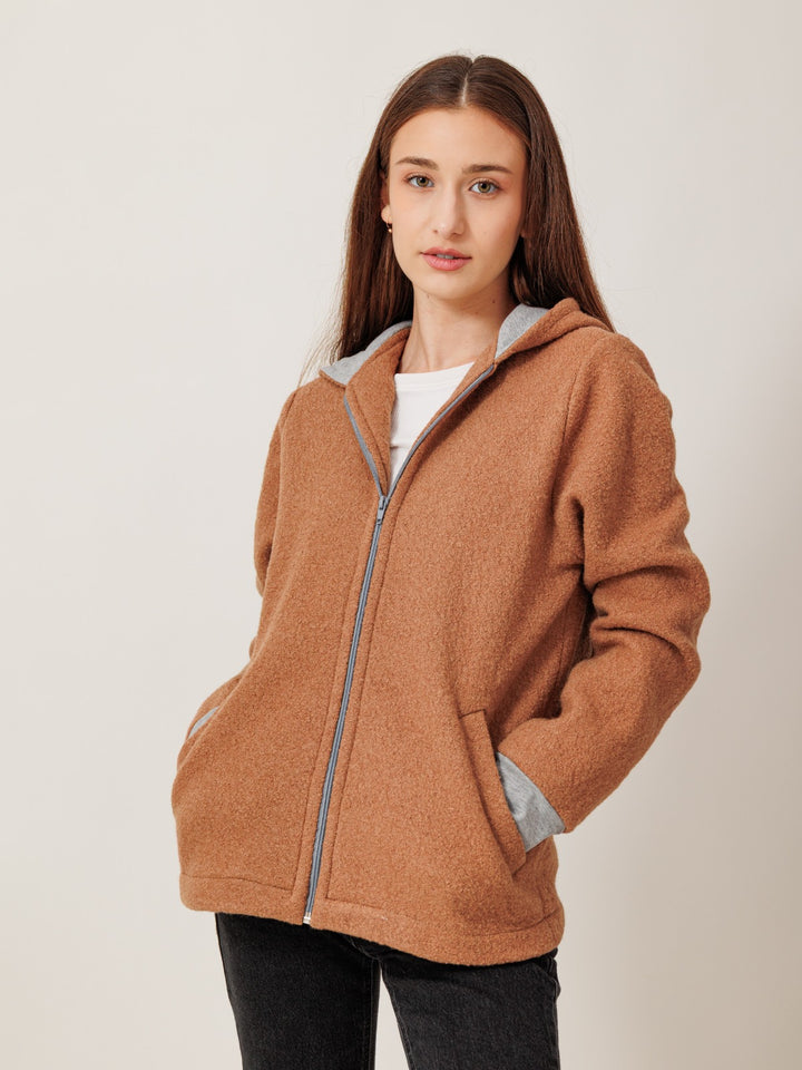 A young woman with long brown hair wearing a brown hooded jacket stands against a plain white background.